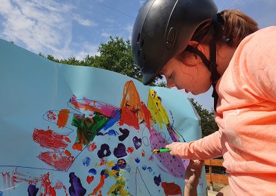 Eine junge Person mit einem Reithelm und einem rosafarbenen Oberteil konzentriert sich darauf, mit einem kleinen Stift auf ein großes, hellblaues Plakat zu malen, das bereits mit bunten, abstrakten Mustern bedeckt ist.