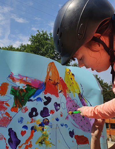 Eine junge Person mit einem Reithelm und einem rosafarbenen Oberteil konzentriert sich darauf, mit einem kleinen Stift auf ein großes, hellblaues Plakat zu malen, das bereits mit bunten, abstrakten Mustern bedeckt ist.