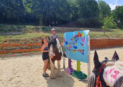 Ein Mädchen mit Helm sitzt auf einem großen, braun-weiß gescheckten Pferd, während eine junge Frau lächelnd das Pferd am Zügel hält. Die Gruppe steht auf einem sandigen Reitplatz neben einer bunten, bemalten Tafel, was auf eine therapeutische oder spielerische Reitaktivität hindeutet.