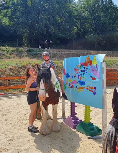 Ein Mädchen mit Helm sitzt auf einem großen, braun-weiß gescheckten Pferd, während eine junge Frau lächelnd das Pferd am Zügel hält. Die Gruppe steht auf einem sandigen Reitplatz neben einer bunten, bemalten Tafel, was auf eine therapeutische oder spielerische Reitaktivität hindeutet.