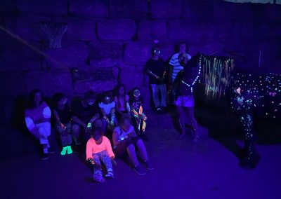 Eine Gruppe von etwa zwölf Menschen, darunter mehrere Kinder, sitzt und steht im Dunkeln vor einer Steinmauer, wobei ihre Kleidung und Körperbemalung durch Schwarzlicht in leuchtenden Neonfarben strahlt.