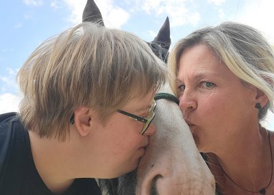 Ein Kind und eine erwachsene Frau küssen gleichzeitig ein Pferd, das im Zentrum des Bildes steht. Das Kind und die Frau neigen sich von links und rechts dem Pferdekopf zu, während sie unter einem blauen Himmel im Freien stehen.