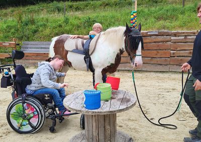 Eine junge Person sitzt in einem Rollstuhl auf einem sandigen Reitplatz neben einem gescheckten Pferd, das von einer weiteren Person auf seinem Rücken geritten wird. Ein Mann hält das Pferd an einer Leine, während das Tier an einem Tisch mit bunten Eimern steht, was auf eine therapeutische Reitstunde hinweist.