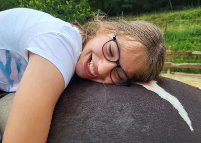 Eine junge Person mit Brille liegt lächelnd und glücklich bäuchlings auf dem Rücken eines dunklen Pferdes und schmiegt sich an dessen Fell. Die Szene, die Vertrauen und Zuneigung zeigt, spielt im Freien auf einem Reitplatz.