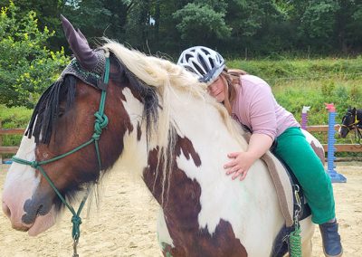 Eine reitende Person mit Helm umarmt fest den Hals eines großen, braun-weiß gescheckten Pferdes mit heller Mähne, wobei sie ihren Kopf liebevoll auflegt. Die Szene spielt auf einem sandigen Reitplatz im Freien, umgeben von grüner Vegetation.