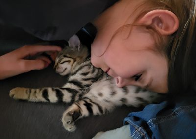 Eine junge Person liegt eng an ein schlafendes, gestromtes Kätzchen geschmiegt, das entspannt auf einer dunklen Decke oder Kleidung liegt.