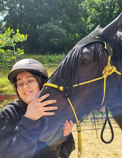 Eine junge Person mit Helm und Brille umarmt liebevoll den Kopf eines schwarzen Pferdes, das eine schwarze Fliegenmaske und ein gelbes Halfter trägt.