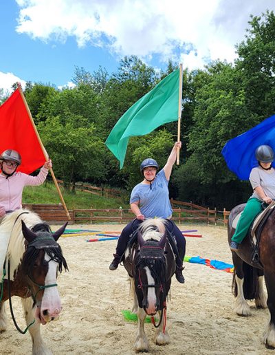 Drei Reiterinnen auf drei Pferden reiten nebeneinander auf einem Sandplatz und schwenken jeweils eine große Fahne