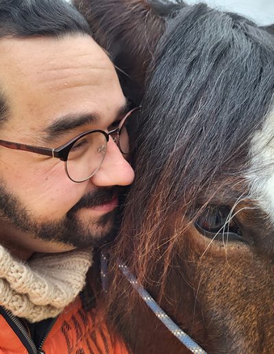 Ein Mann mit Brille und Bart schmiegt seine Wange liebevoll an den Kopf eines braunen Pferdes mit großer weißer Blesse. Es handelt sich um ein Nahaufnahme-Porträt, das die Verbundenheit zwischen dem Mann und dem Pferd zeigt.