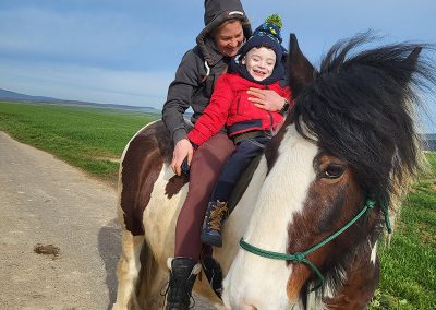 Eine erwachsene Person reitet auf einem großen, braun-weiß gescheckten Pferd und hält ein kleines Kind im roten Anorak vor sich im Sattel. Beide lächeln fröhlich in die Kamera, während das Pferd auf einem Feldweg unter weitem blauem Himmel steht.