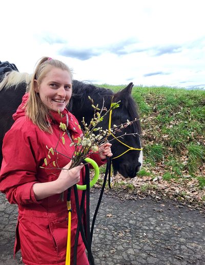 Eine junge Frau in einer leuchtend roten Jacke lächelt in die Kamera und hält einen Strauß blühender Weidenkätzchen. Sie steht neben einem großen, dunklen Pferd mit einem gelben Halfter, das seinen Kopf sanft neben ihr hält.