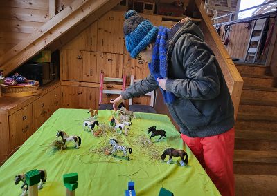 Hier ist die Bildbeschreibung: Eine Person mit blauer Mütze und Schal beugt sich über einen Tisch, auf dem sie eine Szene mit vielen kleinen Spielzeugpferden, Heu und Bauklötzen arrangiert hat. Die Szene findet in einem gemütlichen Raum mit Holzwänden und einer Holztreppe statt.
