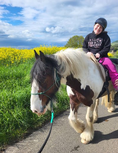 Eine lächelnde Person mit Reithelm reitet auf einem großen, braun-weiß gescheckten Pferd einen Weg entlang.