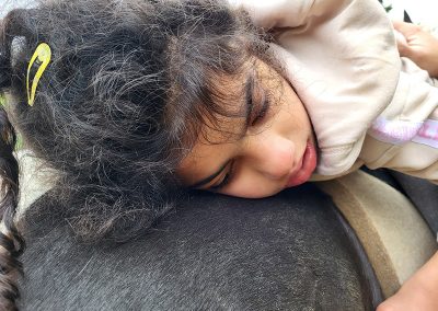Ein Mädchen mit dunklen Locken und einer gelben Haarspange liegt entspannt auf dem dunklen Rücken eines Pferdes. Es trägt einen beigen Kapuzenpullover, und die Szene strahlt Ruhe und Vertrauen aus.