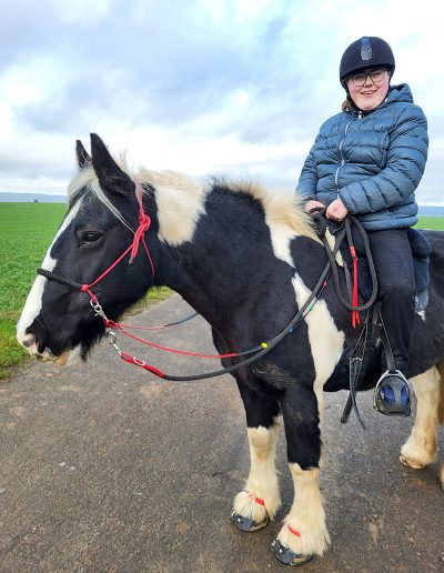 Ein Mädchen mit Helm und Brille sitzt lächelnd auf einem schwarz-weiß gescheckten Pferd.