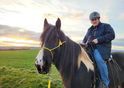 Ein Mann mit Reithelm sitzt auf einem dunklen Pferd mit einer langen Mähne und gelbem Halfter, während sie auf einer grünen Wiese stehen. Das Licht des Sonnenuntergangs oder Sonnenaufgangs erhellt den Himmel im Hintergrund.