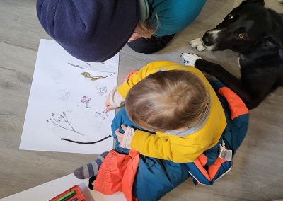 Eine erwachsene Person und ein Kleinkind sitzen gemeinsam auf dem Boden und schauen auf eine Zeichnung, während sie bunte Stifte in einer Schachtel vor sich haben.