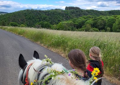 Zwei Kinder laufen neben einem hellen Pferd mit Blumen im Zaumzeug auf einem schmalen Asphaltweg.