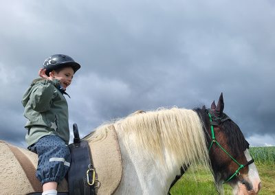 Ein kleines Kind mit Reithelm sitzt lächelnd auf dem Rücken eines braun-weißen Pferdes mit heller Mähne, unter einem dramatisch bewölkten Himmel.