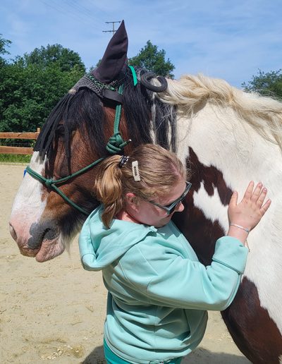 Ein junges Mädchen in einem hellgrünen Kapuzenpullover steht auf einem Sandplatz und umarmt zärtlich den Hals eines braun-weißen Pferdes mit langer Mähne.