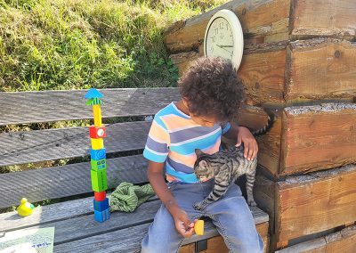 Ein Kind mit lockigem Haar sitzt auf einer Holzbank und streichelt eine grau-getigerte Katze, die auf seinem Schoß steht. Neben dem Kind befindet sich ein hoher Turm aus bunten Bauklötzen.