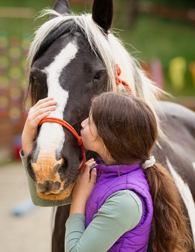 Ein Mädchen mit einem lila Weste und einem Pferdeschwanz umarmt liebevoll den Kopf eines schwarz-weißen Pferdes mit hellweißer Mähne.